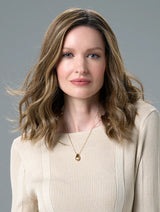 Woman with wavy brown hair wearing a beige sweater and gold necklace against a gray background